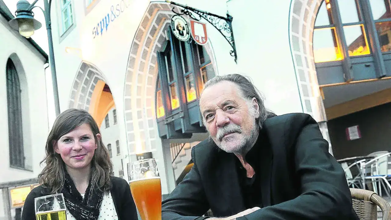 Bernd Schroeder sitzt mit einer jungen Frau bei Bier vor einem historischen Geb&auml;ude.