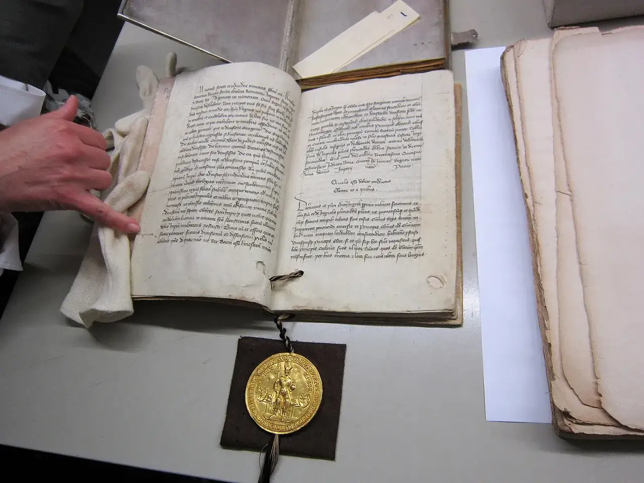 Die goldene Bulle, ein historisches Dokument mit lateinischer Schrift, liegt auf einem Tisch. Eine Hand mit Handschuh wischt vorsichtig über die Seiten.