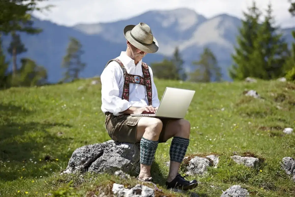 Mann in Lederhose und Hut arbeitet auf einem Laptop in den Bergen.