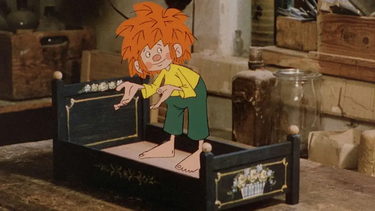 Pumuckl steht in Meister Eders Werkstatt auf einem kleinen Bettchen und schaut neugierig umher.