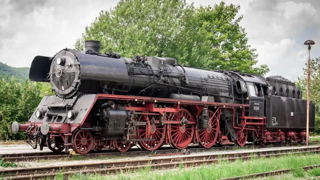 Eine historische Dampflokomotive, ein Symbol der Erfindung Eisenbahn, steht auf Gleisen vor grünen Bäumen.