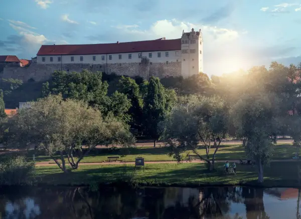 Schloss hinter Bäumen, ein Fluss im Vordergrund. Ein friedlicher Tag, wie ihn die Wettiner heute genießen könnten.