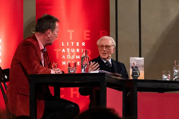 Zwei Männer im Gespräch, einer mit Buch "Franz von Bayern". Einblicke in die bayerische Geschichte, vielleicht aus dem Jahr 794.