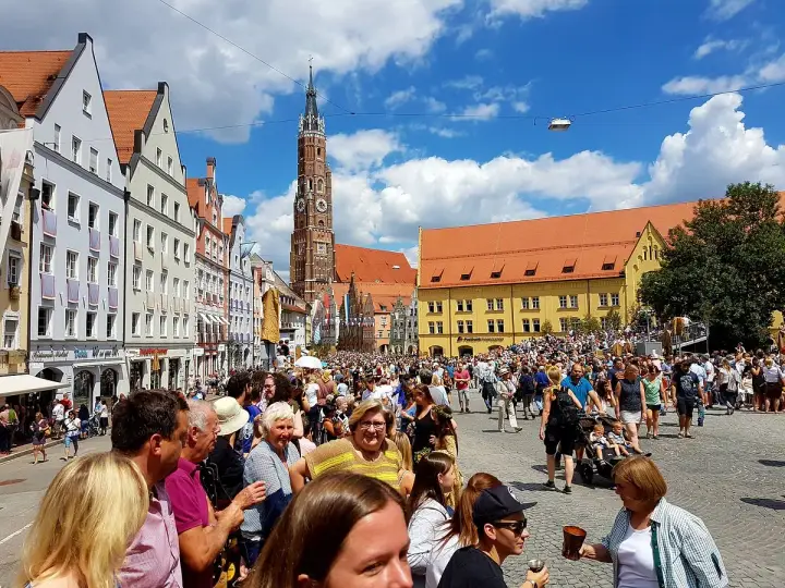 Menschenmassen auf dem Platz der Landshuter Hochzeit, mit historischen Gebäuden und einem Kirchturm im Hintergrund.