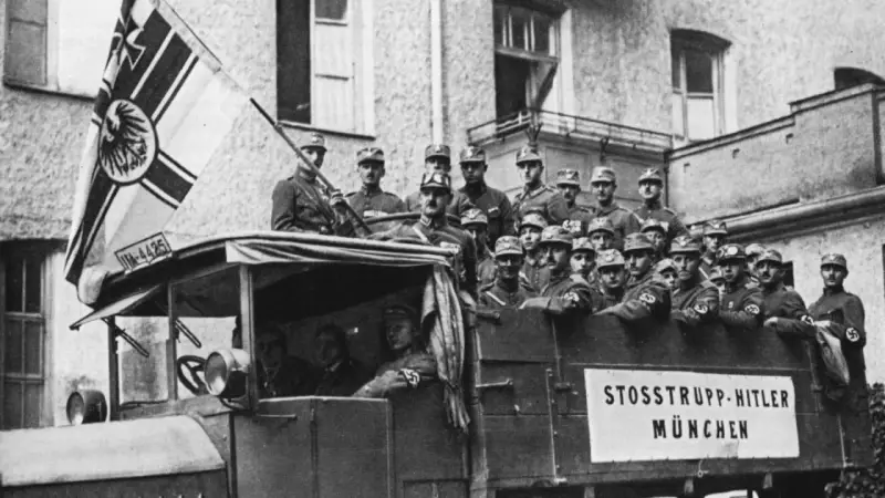 Männer in Uniformen, die an den Hitler-Putsch erinnern, stehen auf einem Lastwagen mit einer Fahne.