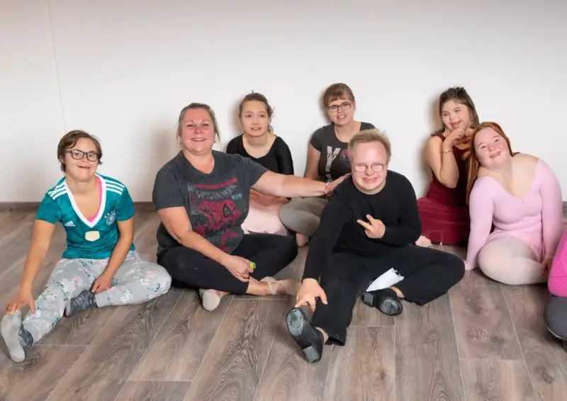 Gruppe von Menschen mit Down-Syndrom und einer Betreuerin sitzen auf dem Boden.